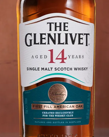 A close-up of a Glenlivet 14 Years Single Malt Scotch Whisky bottle, showing the amber liquid, white and teal label, and gold seal against a warm brown background.