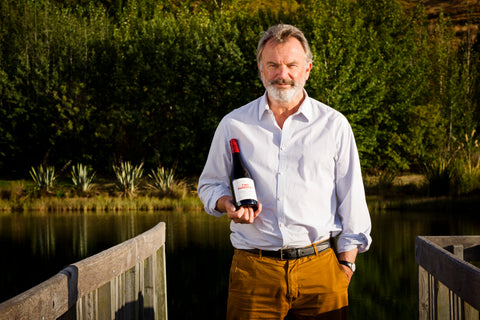Actor Sam Neill stands on a wooden dock holding a bottle of wine, with a lake and green trees in the background.
